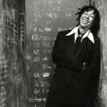 Foto em preto e branco do cantor Djavan sorrindo; ao fundo, uma parede com notas musicais desenhadas a giz